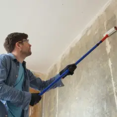Mężczyzna w okularach ochronnych i rękawiczkach maluje ścianę wałkiem.